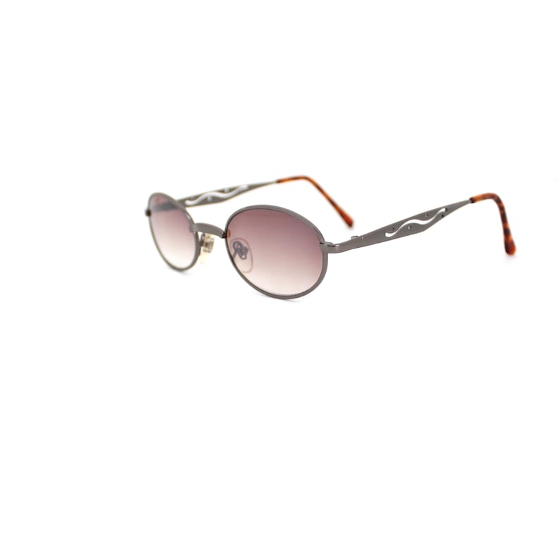 Pode incluir: Um par de &oacute;culos de sol ovais com arma&ccedil;&atilde;o de metal prateado e lentes marrons. A arma&ccedil;&atilde;o tem um design decorativo com uma linha ondulada de cada lado.