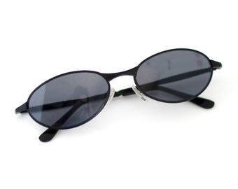 May include: Black oval sunglasses with dark gray lenses. The sunglasses have a thin, black frame and a small bridge. The temples are also black and curve slightly. The sunglasses are set against a white background.