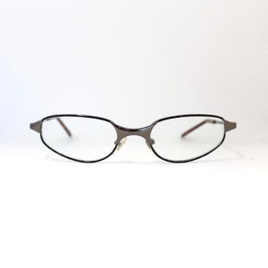 May include: Oval-shaped eyeglasses with a dark brown metal frame and clear lenses. The glasses have a thin, black outline around the lenses and a small nose bridge. The arms of the glasses are also dark brown.