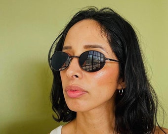 May include: Round black sunglasses with a thin, dark metal frame. The lenses reflect a building's windows. The sunglasses are worn by a person with dark hair and a white tank top. The background is a solid, light green color.