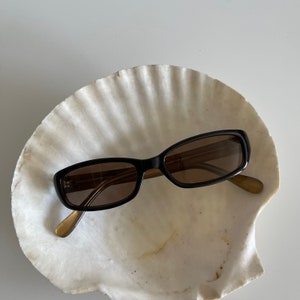 May include: Black sunglasses with brown tinted lenses rest inside a large, white seashell. The sunglasses have a rectangular shape with a black frame and gold-colored arms. The seashell has a textured surface.