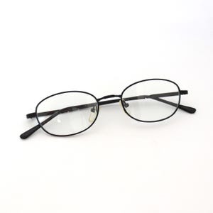 Może przedstawiać: Owalne czarne okulary z przezroczystymi soczewkami. Okulary mają cienką, ciemną oprawkę i zauszniki. Soczewki są czyste i bez zadrapań. Okulary są umieszczone na białym tle.