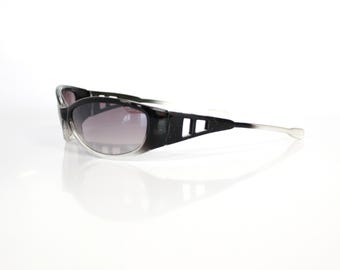 May include: Black and clear sunglasses with a gradient lens. The sunglasses have a thick black frame with rectangular cutouts on the temples and a clear lower frame. The arms are silver.