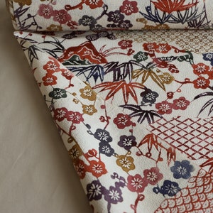 May include: A rolled-up fabric with a white background and a repeating pattern of colorful floral and leaf motifs. The design includes red, purple, and gold flowers, along with green and brown leaves. The fabric has a textured appearance.