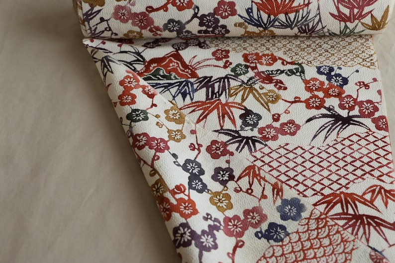 May include: A rolled-up fabric with a white background and a repeating pattern of colorful floral and leaf motifs. The design includes red, purple, and gold flowers, along with green and brown leaves. The fabric has a textured appearance.
