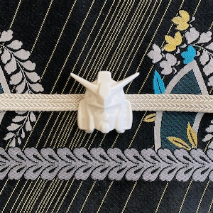 K&ouml;nnte beinhalten: Wei&szlig;e Keramikfigur eines Roboterkopfes mit detailliertem Design. Die Figur ist auf einem wei&szlig;en Stoffband mit einem schwarzen und goldenen Muster im Hintergrund platziert.