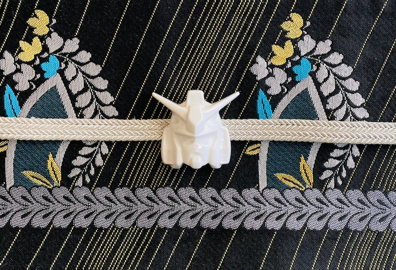 K&ouml;nnte beinhalten: Wei&szlig;e Keramikfigur eines Roboterkopfes mit detailliertem Design. Die Figur ist auf einem wei&szlig;en Stoffband mit einem schwarzen und goldenen Muster im Hintergrund platziert.