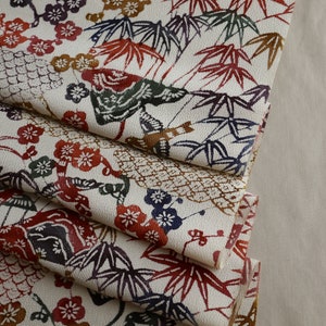 May include: Cream-colored fabric with a repeating pattern of red, brown, blue, and green leaves and flowers. The fabric is folded, revealing layers of the detailed design. The pattern includes bamboo leaves, plum blossoms, and geometric shapes.