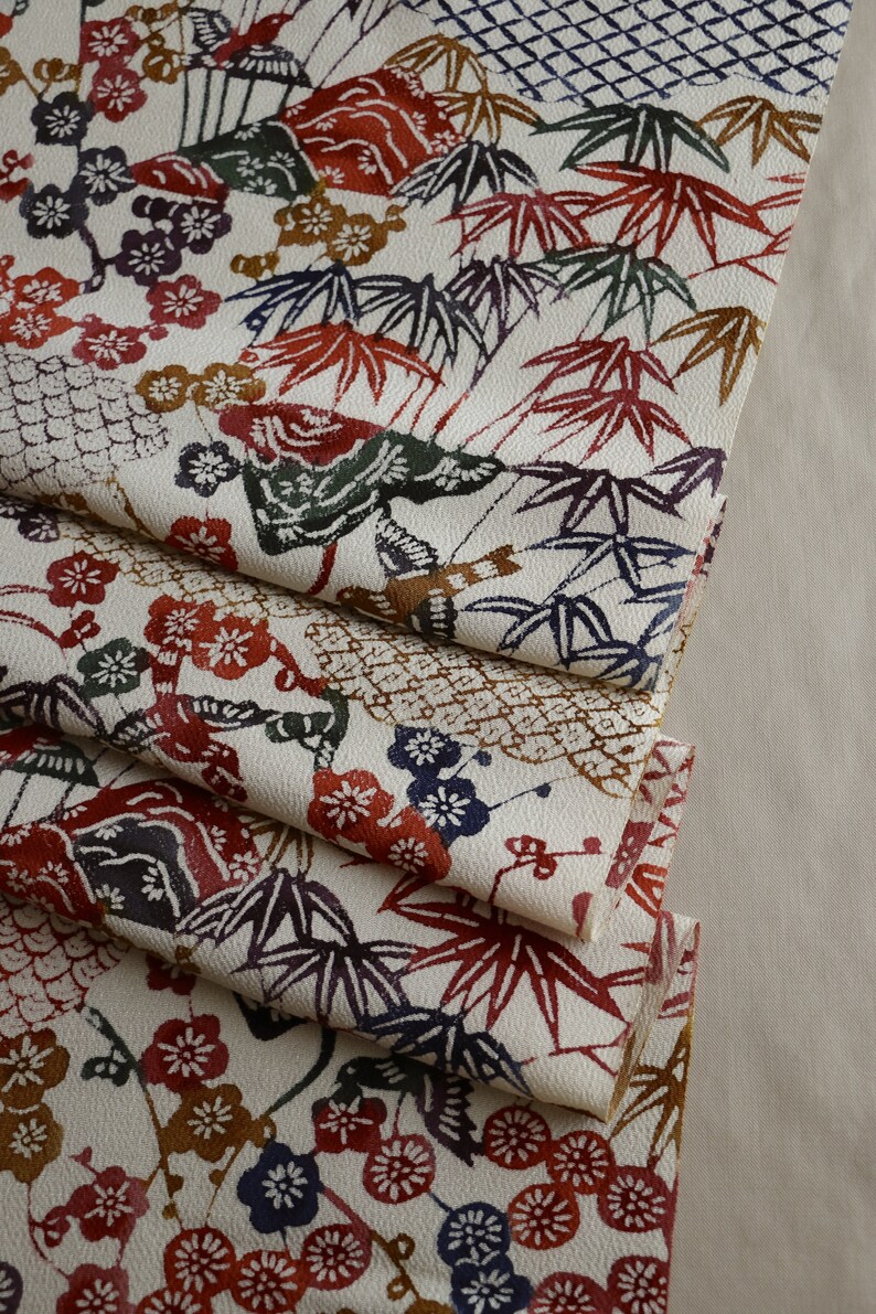 May include: Cream-colored fabric with a repeating pattern of red, brown, blue, and green leaves and flowers. The fabric is folded, revealing layers of the detailed design. The pattern includes bamboo leaves, plum blossoms, and geometric shapes.