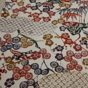 May include: A cream-colored fabric with a repeating pattern of Japanese-style illustrations. The design features red, purple, and blue plum blossoms, bamboo, and birds. The overall aesthetic is reminiscent of traditional Japanese art.