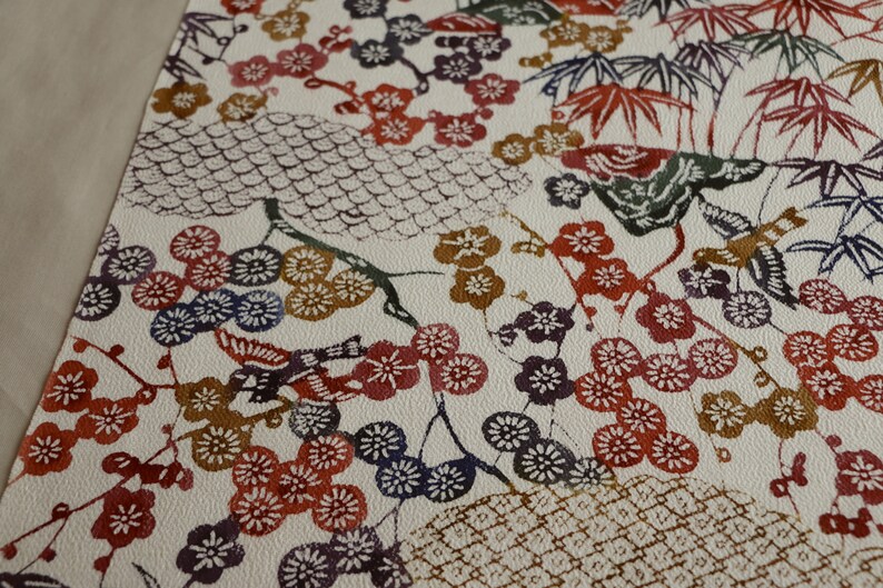May include: A cream-colored fabric with a repeating pattern of Japanese-style illustrations. The design features red, purple, and blue plum blossoms, bamboo, and birds. The overall aesthetic is reminiscent of traditional Japanese art.