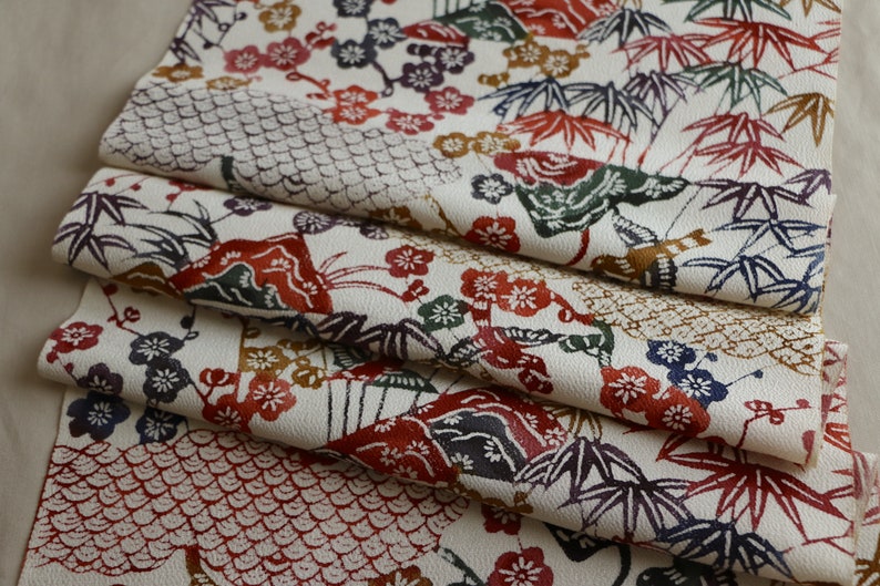 May include: Folded fabric with a cream background and a repeating pattern of Japanese-style motifs. The design includes red, green, and purple bamboo leaves, flowers, and geometric shapes.