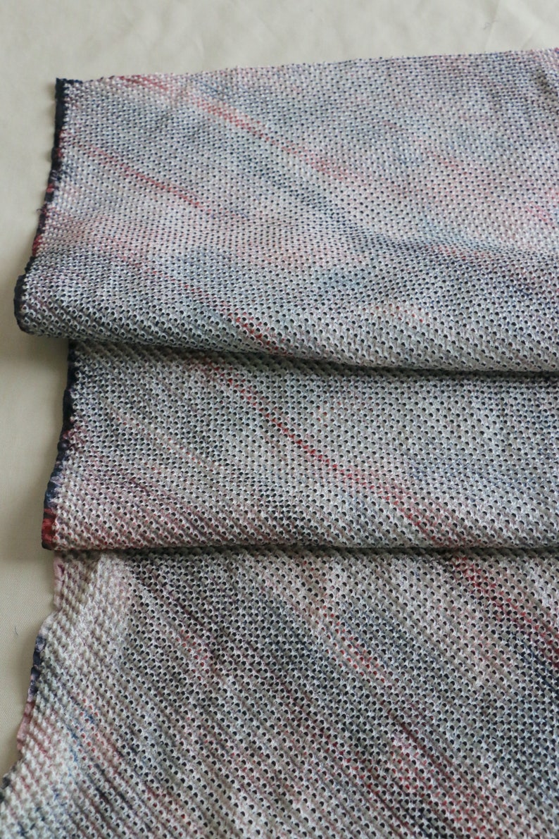 May include: A close-up of a white fabric with a textured pattern of small holes. The fabric has a subtle red and blue striped pattern.