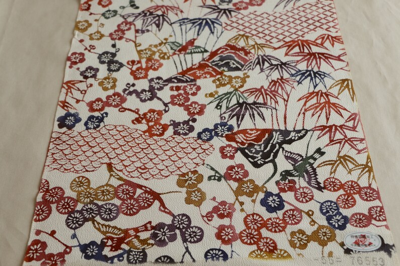 May include: A rectangular piece of paper with a traditional Japanese design. The design features colorful illustrations of flowers, birds, bamboo, and mountains in shades of red, blue, purple, and gold on a white background.