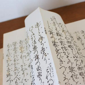 Könnte beinhalten: Ein offenes Buch mit handgeschriebener japanischer Kalligraphie in schwarzer Tinte auf gealtertem, cremefarbenem Papier. Die Seiten sind leicht gewölbt und zeigen den Text und die Textur des Papiers. Das Buch liegt auf einer Holzoberfläche.