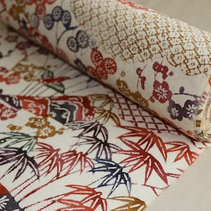 May include: Rolled fabric with a detailed floral and geometric pattern. The design features red, brown, and purple flowers and bamboo leaves on a cream background. The fabric is rolled up, revealing the layered edges.