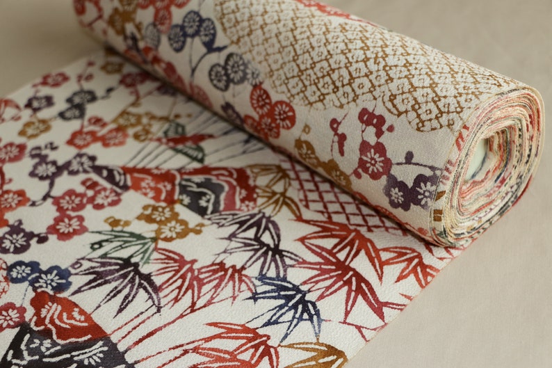 May include: Rolled fabric with a detailed floral and geometric pattern. The design features red, brown, and purple flowers and bamboo leaves on a cream background. The fabric is rolled up, revealing the layered edges.