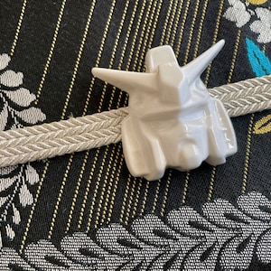 K&ouml;nnte beinhalten: Wei&szlig;e Keramikfigur eines Roboterkopfes mit detailliertem Design. Die Figur ist auf einem beigefarbenen geflochtenen Band vor einem schwarzen und goldenen gemusterten Stoff platziert.