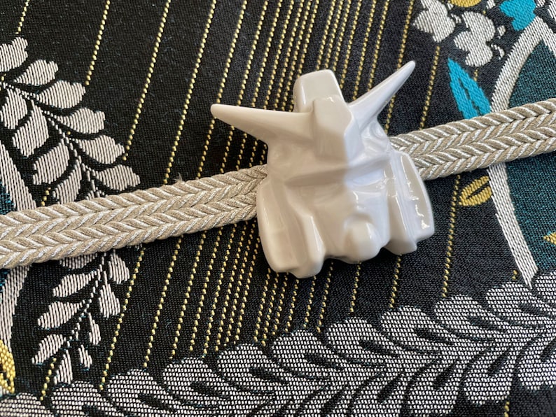 K&ouml;nnte beinhalten: Wei&szlig;e Keramikfigur eines Roboterkopfes mit detailliertem Design. Die Figur ist auf einem beigefarbenen geflochtenen Band vor einem schwarzen und goldenen gemusterten Stoff platziert.