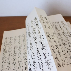 Könnte beinhalten: Ein offenes Buch mit handschriftlichem japanischem Text in schwarzer Tinte auf weißem Papier. Die Seiten sind umgeklappt und das Buch liegt auf einer Holzoberfläche.