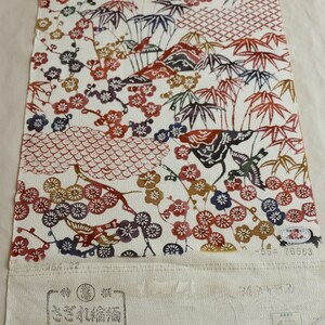 May include: A rolled-up textile with a white background, featuring a repeating pattern of plum blossoms, bamboo, and birds in shades of red, brown, blue, and gold. The fabric has Japanese text and stamps.