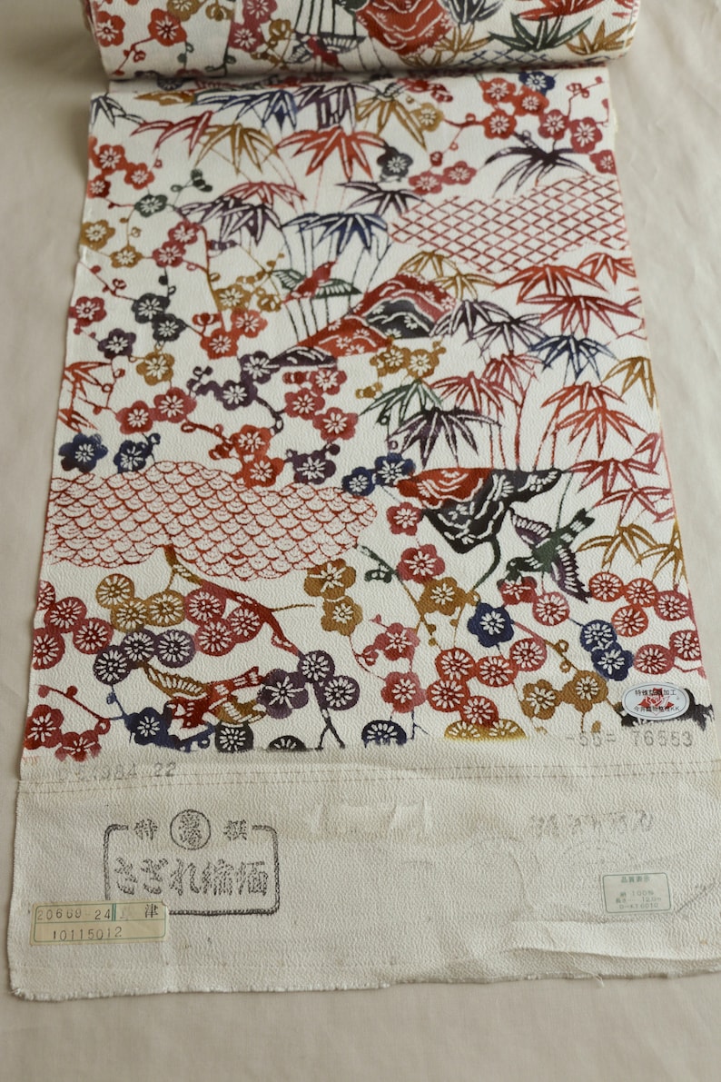 May include: A rolled-up textile with a white background, featuring a repeating pattern of plum blossoms, bamboo, and birds in shades of red, brown, blue, and gold. The fabric has Japanese text and stamps.