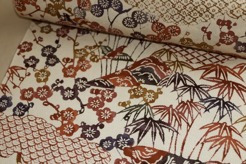 May include: Cream-colored fabric with a repeating pattern of Japanese-style illustrations. The design features plum blossoms, bamboo, and stylized mountains in shades of red, brown, purple, and green. The fabric is likely used for clothing or home decor.