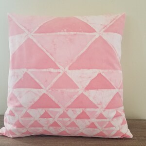 May include: A square decorative pillow with a geometric pattern. The pillow features a pink background with white triangular shapes arranged in rows. The pillow is resting on a wooden surface.