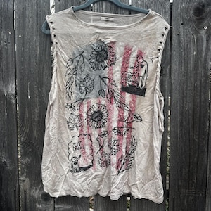 May include: Beige sleeveless shirt with a graphic design featuring sunflowers, leaves, and red stripes. The shirt has cut-out details on the shoulders and the text "WHAT CAN I LEARN?" is visible.