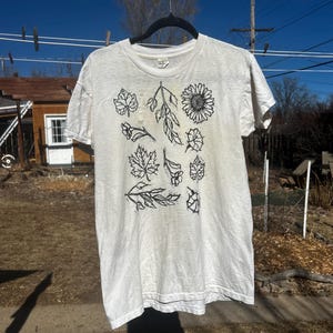 May include: A white t-shirt with a black floral design, including leaves and a sunflower. The shirt is hanging on a clothesline with clothespins against a blue sky.