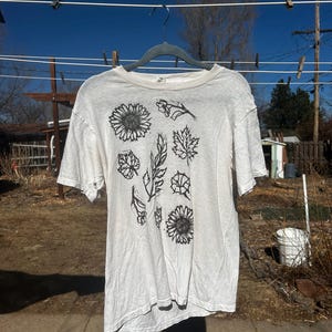 May include: White t-shirt with a crew neck and short sleeves. The shirt features a black floral design, including sunflowers and leaves. The t-shirt is hanging on a clothesline against a bright blue sky.