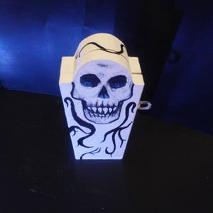 Puede incluir: Una caja de madera blanca con forma de ataúd con un diseño de calavera y vid en blanco y negro pintado en la parte delantera.