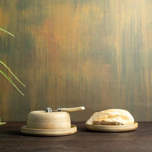 Könnte beinhalten: Eine hellbraune Keramik-Butterdose mit Deckel und Buttermesser, daneben ein Teller mit geschnittenem Brot. Das Messer hat einen hellfarbigen Griff. Der Hintergrund ist eine strukturierte Wand mit grünen, braunen und orangenen Tönen.