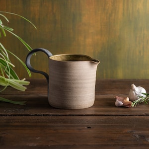 Könnte beinhalten: Handgefertigter Keramik-Krug mit dunklem Griff und Ausguss. Der Krug hat eine strukturierte, hellbraune Außenseite und eine dunkle Innenseite. Der Hintergrund umfasst Grün, Knoblauch und Rosmarin auf einer Holzoberfläche.