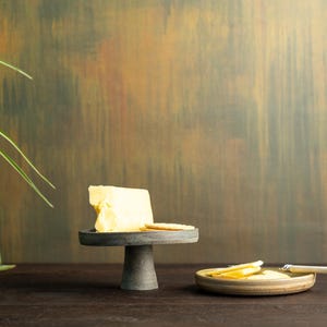 Könnte beinhalten: Ein Stillleben mit einem Block gelber Butter auf einem kleinen, dunkelgrauen Sockel. Cracker sind auf dem Sockel und einem kleinen Teller platziert. Ein Buttermesser liegt auf dem Teller mit geschnittener Butter. Der Hintergrund ist eine strukturierte, mehrfarbige Wand.