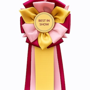 May include: A rosette ribbon featuring layered satin fabric in deep burgundy, soft pink, and golden yellow. A central circular gold disc displays the text BEST IN SHOW in dark red lettering. Three long vertical ribbons hang from the base.