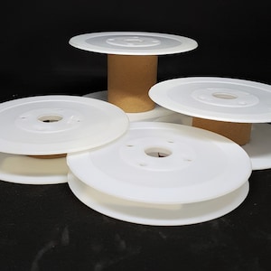 Op de afbeelding: Verschillende witte plastic spoelen met een centraal gat en een kartonnen kern zijn geplaatst tegen een zwarte achtergrond. De spoelen zijn cirkelvormig met een plat, schijfachtig ontwerp en zijn in een aantrekkelijke opstelling gerangschikt.