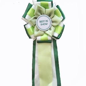 May include: A rosette award ribbon featuring layered satin fabric in shades of dark green, lime green, and white. The center circular disc is white with a dark green border and the text BEST IN SHOW printed in dark green capital letters.
