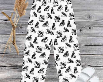 May include: White pajama pants with a black raven pattern. The ravens are perched on rocks and branches. The pants are made of a soft material and are suitable for lounging. White sandals are next to the pants.