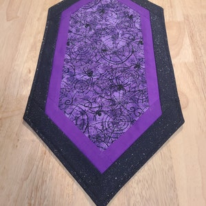May include: A Halloween-themed table runner with a diamond shape. The center features a purple fabric with black spiderwebs and spiders. It is framed by a solid purple border and a black glittery border. The runner is placed on a wooden surface.