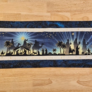 May include: A blue and gold Christmas table runner with a silhouette of the nativity scene and the three wise men. The scene is set against a starry night sky.