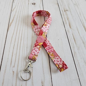 May include: A pink and red patterned lanyard with a silver-colored clasp and key ring. The fabric features a patchwork design with floral and geometric patterns in shades of pink, red, and gold. The lanyard is designed to be worn around the neck.