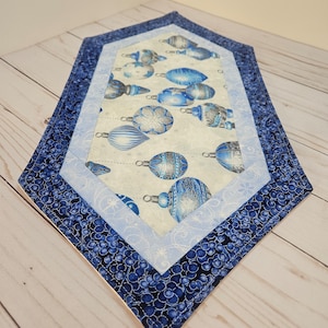 May include: A blue and white quilted table runner with a Christmas ornament design. The runner has a diamond shape with a white background and blue trim.