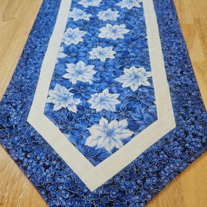 May include: A blue and white floral table runner. The runner features a central panel with white poinsettia flowers on a blue background, framed by a white border. The outer edges have a pattern of blue berries.