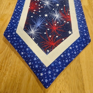 May include: A patriotic table runner with a navy blue background featuring white stars and a central panel with red, white, and blue fireworks. The runner has a white border and a blue star-patterned edge.