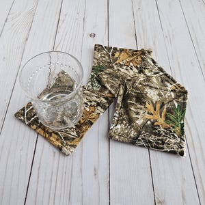 May include: A clear glass sits on a camouflage-patterned square coaster. Additional coasters with the same pattern are visible. The coasters feature a mix of brown, green, and tan colors, resembling a woodland design. The coasters are likely made of fabric.