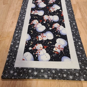 May include: A black and white table runner with a white border and a snowflake pattern. The center of the runner features a repeating pattern of snowmen with red scarves and hats on a black background.