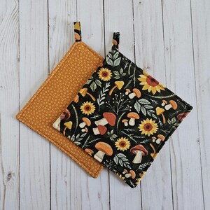 May include: Two square pot holders. One is mustard yellow with an X and O pattern. The other is black with a pattern of sunflowers, mushrooms, and foliage. Both have a small loop for hanging.