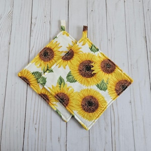 May include: Two white pot holders with a sunflower pattern. The pot holders have a loop for hanging.