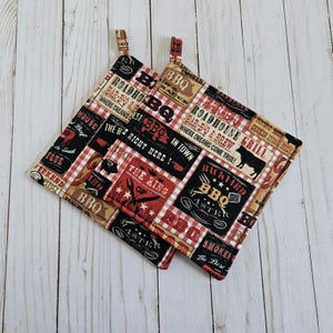 May include: Two black and white pot holders with red and white checkered fabric. The pot holders have a BBQ theme with text that reads "Roadhouse Grill", "Backyard BBQ", "The King of BBQ", and "Smoked BBQ".